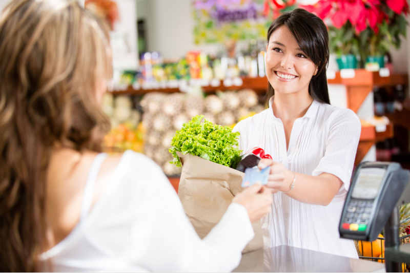 A cashier smiles while handing a grocery bag to a customer at a checkout counter.