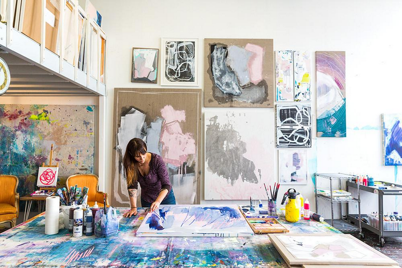 An artist works on a painting in a bright studio filled with abstract artworks.