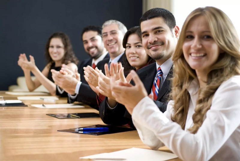 A group of people in business attire sitting at a conference table and clapping.