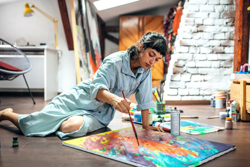 A person painting a colorful artwork on a canvas placed on the floor of an art studio.