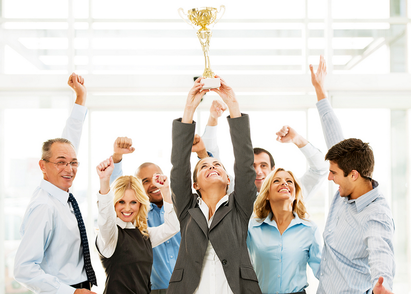 A group of jubilant business professionals celebrates as one woman holds up a trophy.